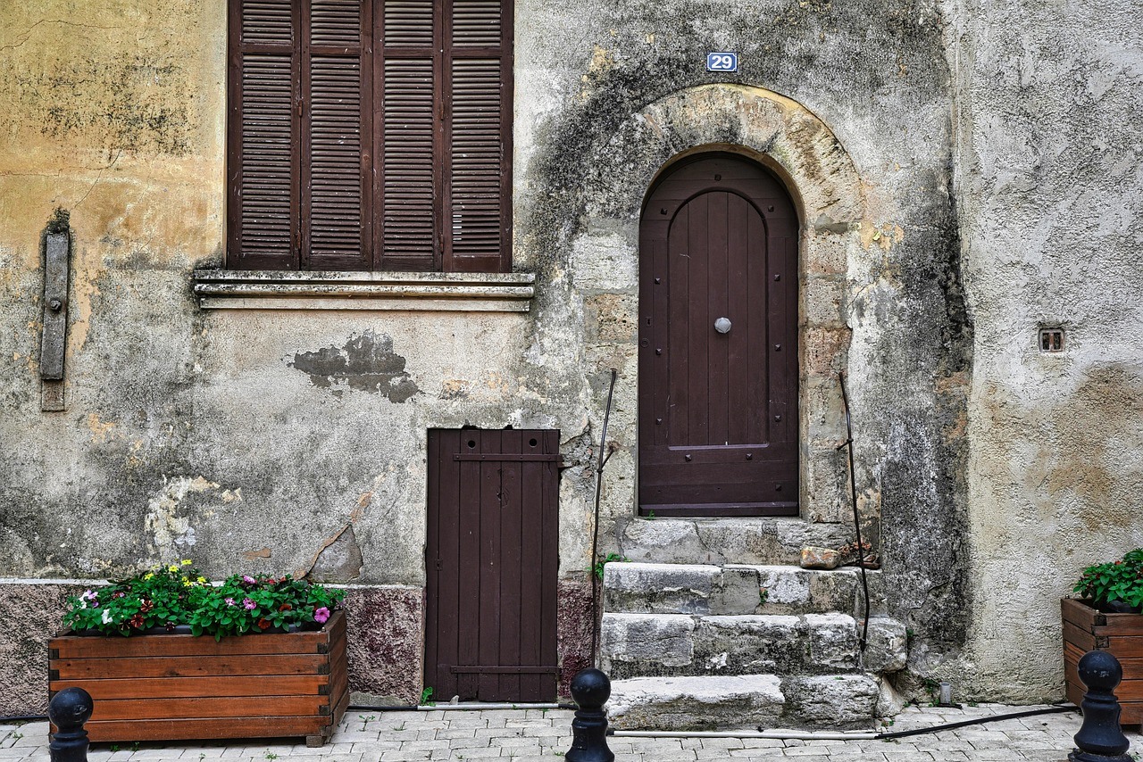 A weathered stone façade contains closed wooden shutters, an arched upper door, a smaller lower door and flower boxes.