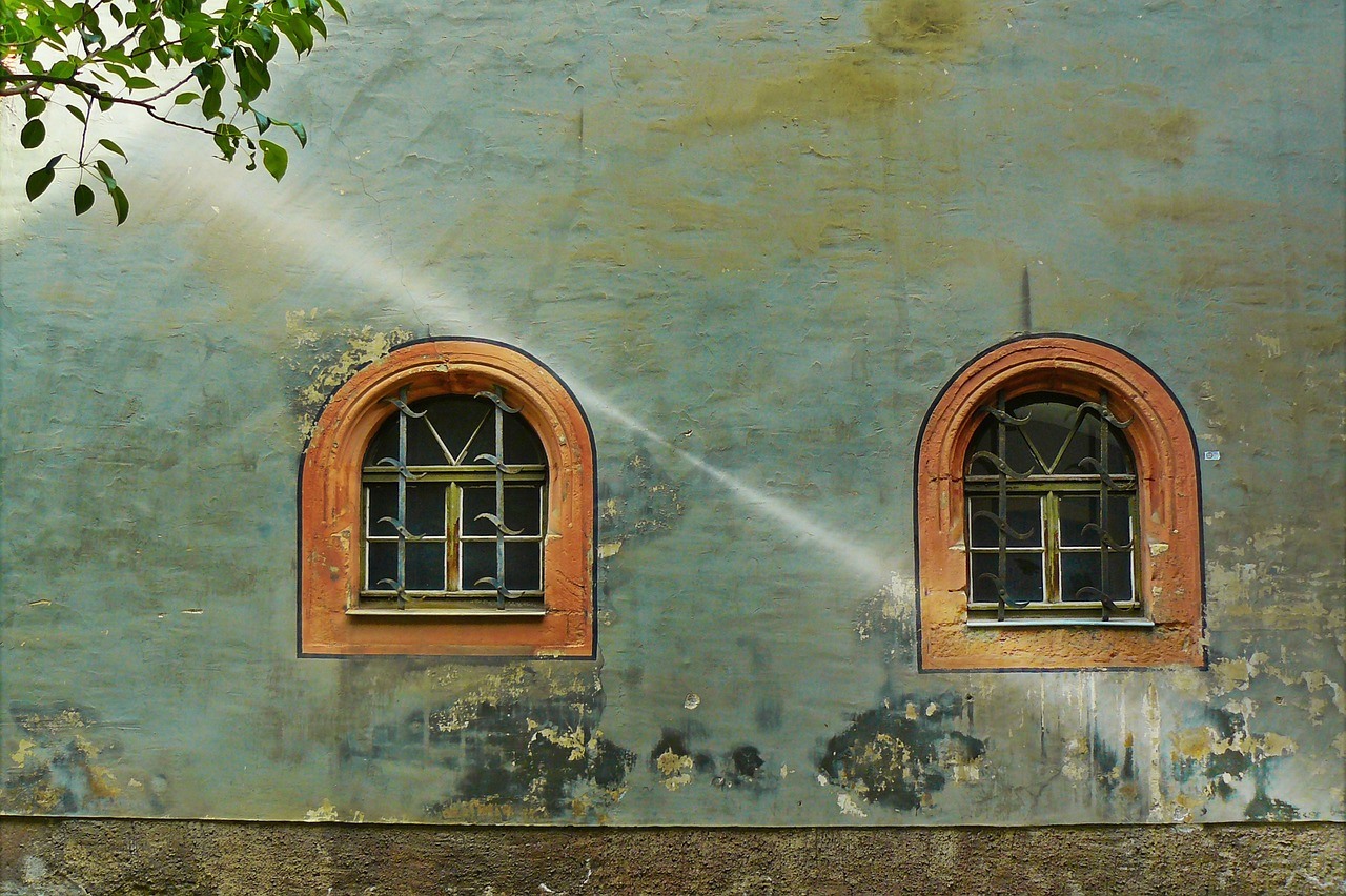 Two small arched windows with orange surrounds sit in a faded blue-green wall crossed by diagonal sunbeams.